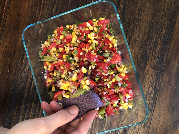 A hand reaching into a clear bowl of salsa